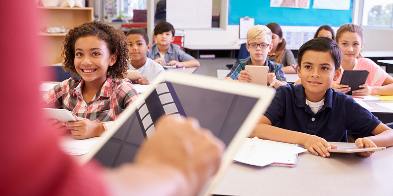 Children in classroom setting looking towards teacher holding a tablet for digital learning